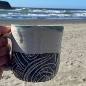 May include: A ceramic mug featuring a two-tone design with a smooth white upper section and a dark blue lower section decorated with carved, swirling white line patterns. A light blue glaze drip runs down from the rim onto the white surface.