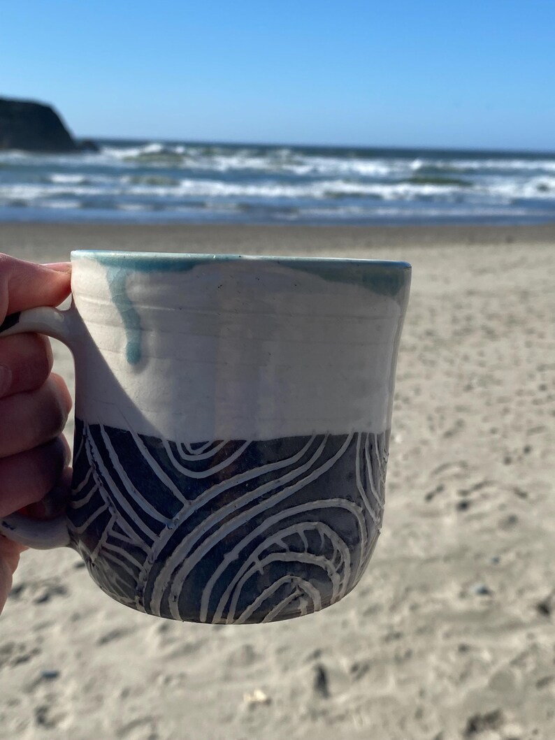 May include: A ceramic mug featuring a two-tone design with a smooth white upper section and a dark blue lower section decorated with carved, swirling white line patterns. A light blue glaze drip runs down from the rim onto the white surface.