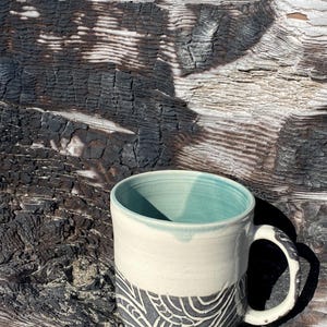 May include: A ceramic mug featuring a smooth cream-colored upper section and a textured dark gray lower section with a carved white swirling line pattern. The interior of the mug is glazed in a soft mint green, and it includes a curved handle.