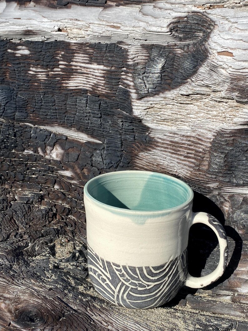 May include: A ceramic mug featuring a smooth cream-colored upper section and a textured dark gray lower section with a carved white swirling line pattern. The interior of the mug is glazed in a soft mint green, and it includes a curved handle.