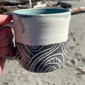 May include: A ceramic mug featuring a two-tone design with a smooth white upper section and a dark gray lower section. The lower portion is decorated with carved, swirling white line patterns. The interior of the mug is a light teal color.