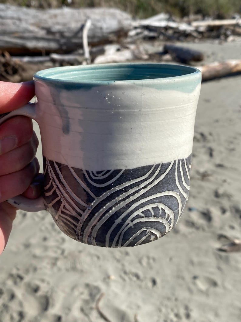 May include: A ceramic mug featuring a two-tone design with a smooth white upper section and a dark gray lower section. The lower portion is decorated with carved, swirling white line patterns. The interior of the mug is a light teal color.