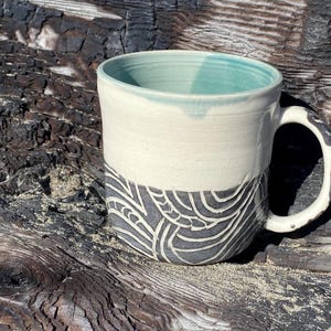 May include: A ceramic mug featuring a smooth cream-colored upper section and a dark gray lower section decorated with carved, swirling white line patterns. The interior of the mug is glazed in a soft teal color, and the handle has a dark, textured finish.