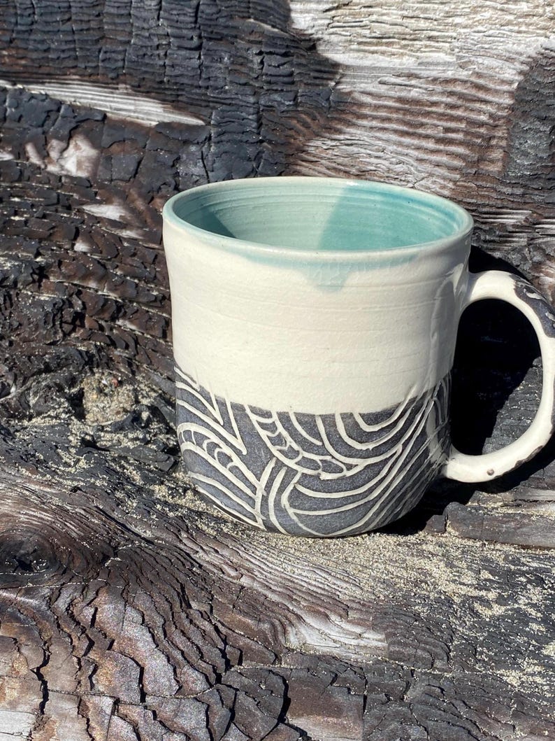 May include: A ceramic mug featuring a smooth cream-colored upper section and a dark gray lower section decorated with carved, swirling white line patterns. The interior of the mug is glazed in a soft teal color, and the handle has a dark, textured finish.