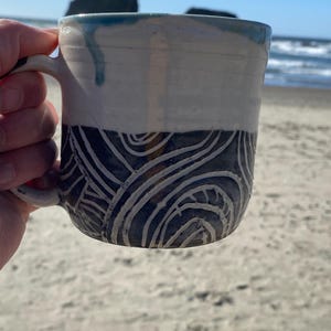 May include: A ceramic mug featuring a two-tone design with a smooth, off-white upper section and a dark gray lower section. The lower portion is decorated with carved, swirling white line patterns. A blue glaze drip accents the top rim.
