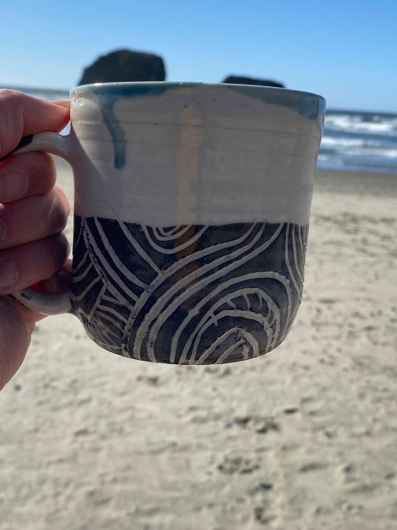 May include: A ceramic mug featuring a two-tone design with a smooth, off-white upper section and a dark gray lower section. The lower portion is decorated with carved, swirling white line patterns. A blue glaze drip accents the top rim.