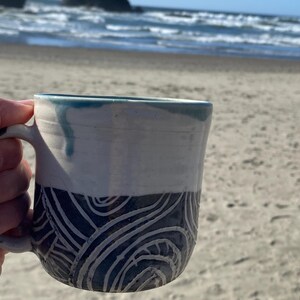May include: A ceramic mug featuring a two-tone design with a smooth white upper section and a dark blue lower section decorated with carved, swirling white line patterns. The rim has a subtle blue glaze drip detail and a curved handle on the side.