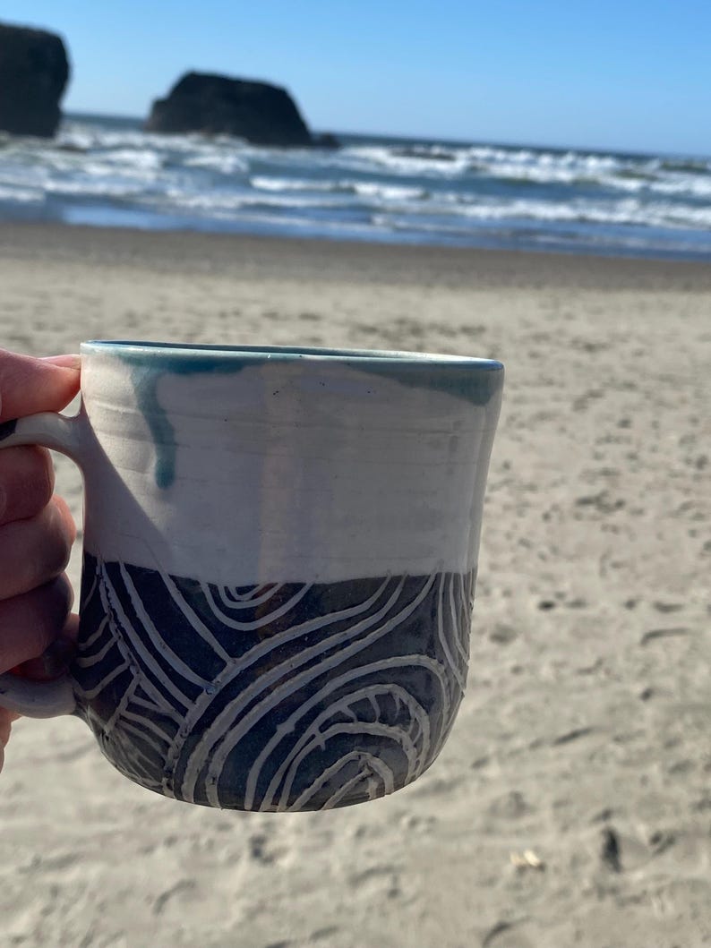May include: A ceramic mug featuring a two-tone design with a smooth white upper section and a dark blue lower section decorated with carved, swirling white line patterns. The rim has a subtle blue glaze drip detail and a curved handle on the side.