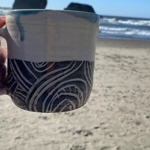 May include: A ceramic mug featuring a two-tone design with a smooth white upper section and a dark lower section decorated with carved, swirling white line patterns. The rim has a subtle blue glaze drip, and the mug has a curved handle on the side.