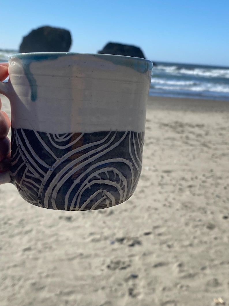 May include: A ceramic mug featuring a two-tone design with a smooth white upper section and a dark lower section decorated with carved, swirling white line patterns. The rim has a subtle blue glaze drip, and the mug has a curved handle on the side.