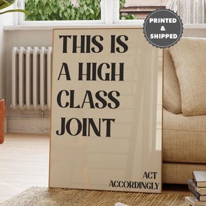 May include: A framed poster with the text "THIS IS A HIGH CLASS JOINT" in black, bold letters. Below the text is "ACT ACCORDINGLY". The poster is displayed in a living room setting with a beige sofa, a potted plant, and a window.