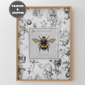 May include: Framed print with a vintage bee postage stamp design. The stamp, valued at 5 cents, displays a detailed bee illustration. The background is a black and white floral pattern, enclosed in a wooden frame.