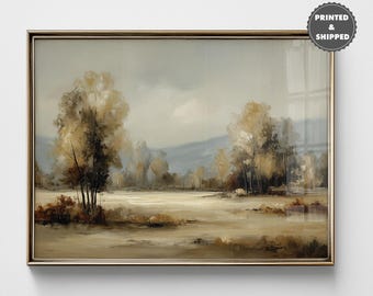 May include: A framed landscape painting with a gold-colored frame. The artwork depicts a serene scene with trees, a field, and a mountain range in the background. The color palette includes muted tones of beige, brown, and gray.