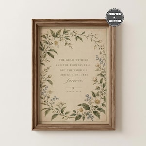May include: A framed wall art print featuring a beige background with a floral wreath border in shades of green, white, and blue. The text reads: The grass withers and the flowers fall, but the word of our God endures forever. Isaiah 40:8.
