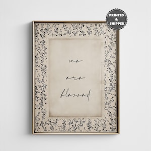 Puede incluir: Arte de pared enmarcado con un marco dorado. La obra presenta las palabras "we are blessed" en una fuente manuscrita sobre un fondo crema. El marco tiene un patrón floral negro.