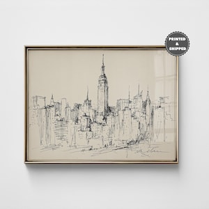 Puede incluir: Una lámina de arte enmarcada con un boceto en tinta negra del horizonte de Nueva York sobre un fondo beige. El dibujo destaca el Empire State Building y los rascacielos circundantes. Una insignia circular en la esquina dice PRINTED & SHIPPED.