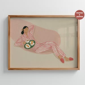 May include: A framed art print shows a person in red and white striped pyjamas, relaxing with a plate of eggs and coffee. The illustration has a soft, pastel colour palette and is set against a neutral background.