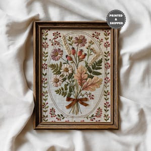 May include: A rectangular wooden frame containing an embroidery of a floral bouquet featuring pink flowers, wheat stalks, green ferns, and oak leaves, tied with a brown ribbon. The design is enclosed in an oval border with small red berries.