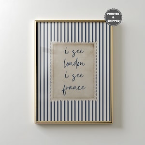 May include: Framed art print with a gold-colored frame. The artwork features a vintage-style postage stamp design with the words "i see london i see france" in a handwritten font. The background has navy blue and white vertical stripes.