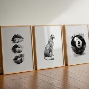 May include: Three framed art prints leaning against a wall. The prints feature black and white images: three sets of lips, a leopard, and a pool ball with the number 8 and the text "LUCKY ME". The frames are light wood.