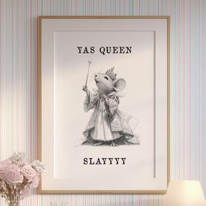 May include: A framed print with a gold border featuring a mouse dressed in regal attire, holding a wand. The text "YAS QUEEN" and "SLAYYYY" are above and below the mouse illustration. The print is displayed on a white desk with a lamp and flowers.