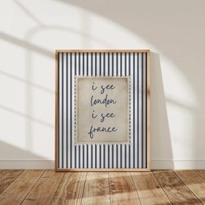 May include: A framed print with a navy blue and white striped background. The print features a central rectangular design with the text "i see london i see france" in a handwritten style. The frame is a light brown color.