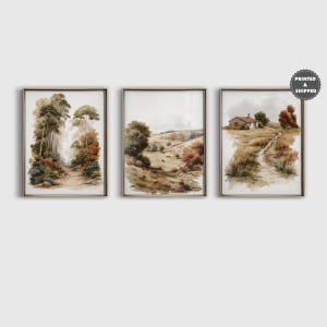 May include: A set of three framed landscape art prints featuring watercolour-style scenes. The left print shows a forest path, the centre depicts rolling hills, and the right displays a small cottage with a dirt path, all in muted earth tones.
