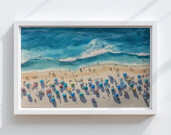 Könnte beinhalten: Gerahmtes Aquarellgemälde, das eine sonnige Strandszene zeigt. Türkisfarbene Wellen branden an den sandigen Ufern, wo bunte Sonnenschirme die Landschaft säumen. Menschen verteilen sich am Strand. Das Gemälde ist in einem weißen Rahmen.