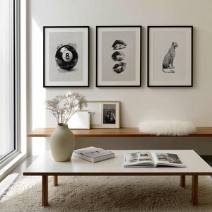 May include: Three framed black and white prints hang on a white wall. The prints depict a pool ball with the number 8, three sets of lips, and a leopard. A white coffee table with a book and vase is in front.