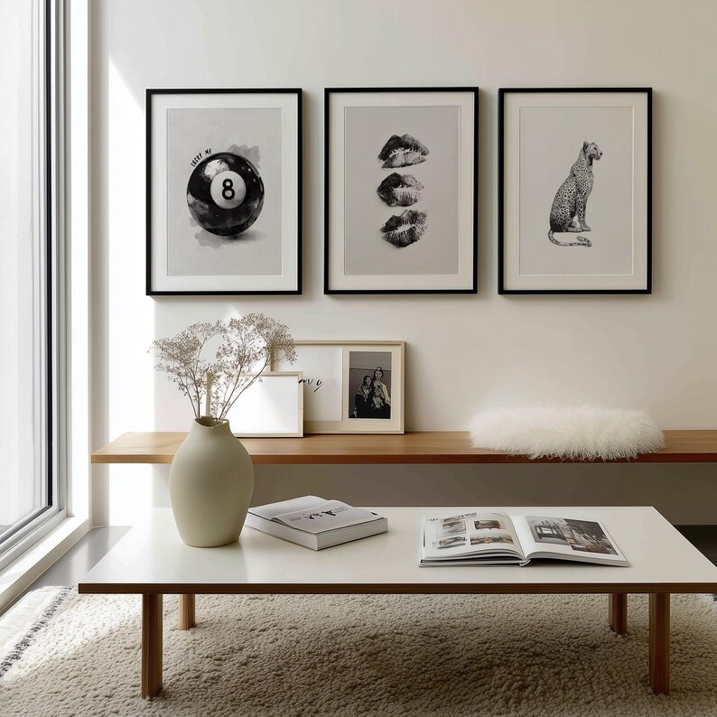 May include: Three framed black and white prints hang on a white wall. The prints depict a pool ball with the number 8, three sets of lips, and a leopard. A white coffee table with a book and vase is in front.