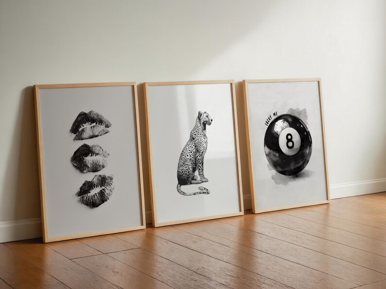 May include: Three framed art prints leaning against a wall. The prints feature black and white images: three sets of lips, a leopard, and a pool ball with the number 8 and the text "LUCKY ME". The frames are light wood.