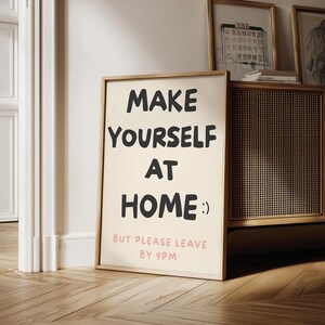 K&ouml;nnte beinhalten: Gerahmtes Poster mit dem Text "MAKE YOURSELF AT HOME :) BUT PLEASE LEAVE BY 9PM" in Schwarz und Rosa. Das Poster befindet sich in einem hellen Holzrahmen und lehnt an einer Wand. Der Hintergrund umfasst einen Holzschrank und gerahmte Kunstwerke.