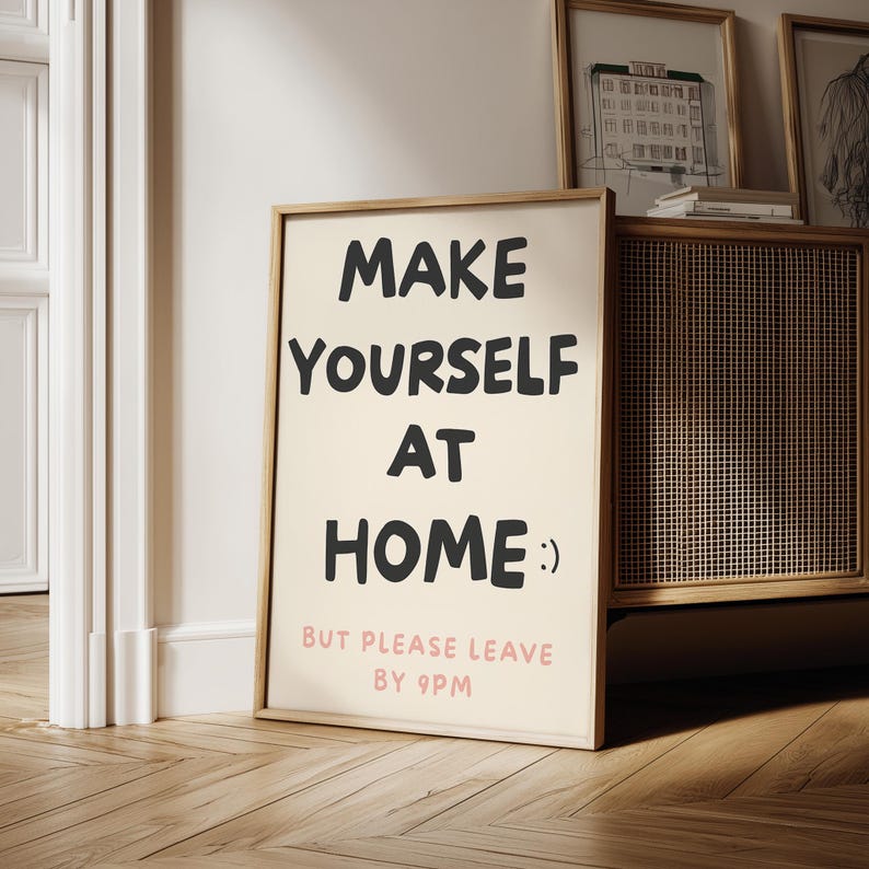 K&ouml;nnte beinhalten: Gerahmtes Poster mit dem Text "MAKE YOURSELF AT HOME :) BUT PLEASE LEAVE BY 9PM" in Schwarz und Rosa. Das Poster befindet sich in einem hellen Holzrahmen und lehnt an einer Wand. Der Hintergrund umfasst einen Holzschrank und gerahmte Kunstwerke.
