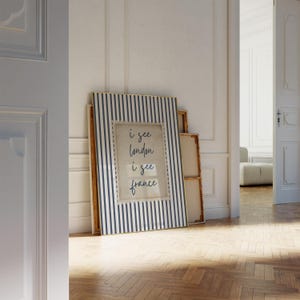 May include: A framed print with a blue and white striped border, leaning against a wall. The print features the text "i see london i see france" in a handwritten style. Other unframed canvases are stacked behind it.