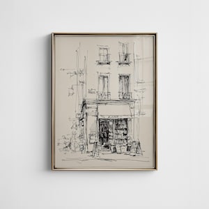Op de afbeelding: Ingelijste zwart-wit schets van een gebouw met een boekwinkel. Het kunstwerk toont een gedetailleerde gevel met ramen, een luifel en een bord met de tekst "BOOKS". De schets zit in een goudkleurige lijst.