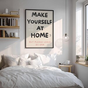 K&ouml;nnte beinhalten: Ein Schlafzimmer mit einem Bett, das mit wei&szlig;er Bettw&auml;sche und Kissen bedeckt ist. Ein gerahmtes Poster an der Wand zeigt den Text "MAKE YOURSELF AT HOME :) BUT PLEASE LEAVE BY 9PM." Ein kleiner Nachttisch aus Holz und eine H&auml;ngeleuchte sind ebenfalls zu sehen.