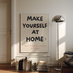K&ouml;nnte beinhalten: Gerahmtes Poster mit dem Text "MAKE YOURSELF AT HOME :) BUT PLEASE LEAVE BY 9PM" in Schwarz und Rosa. Das Poster h&auml;ngt an einer wei&szlig;en Wand &uuml;ber einem Glasregal mit B&uuml;chern. Rechts steht eine silberne Stehlampe.