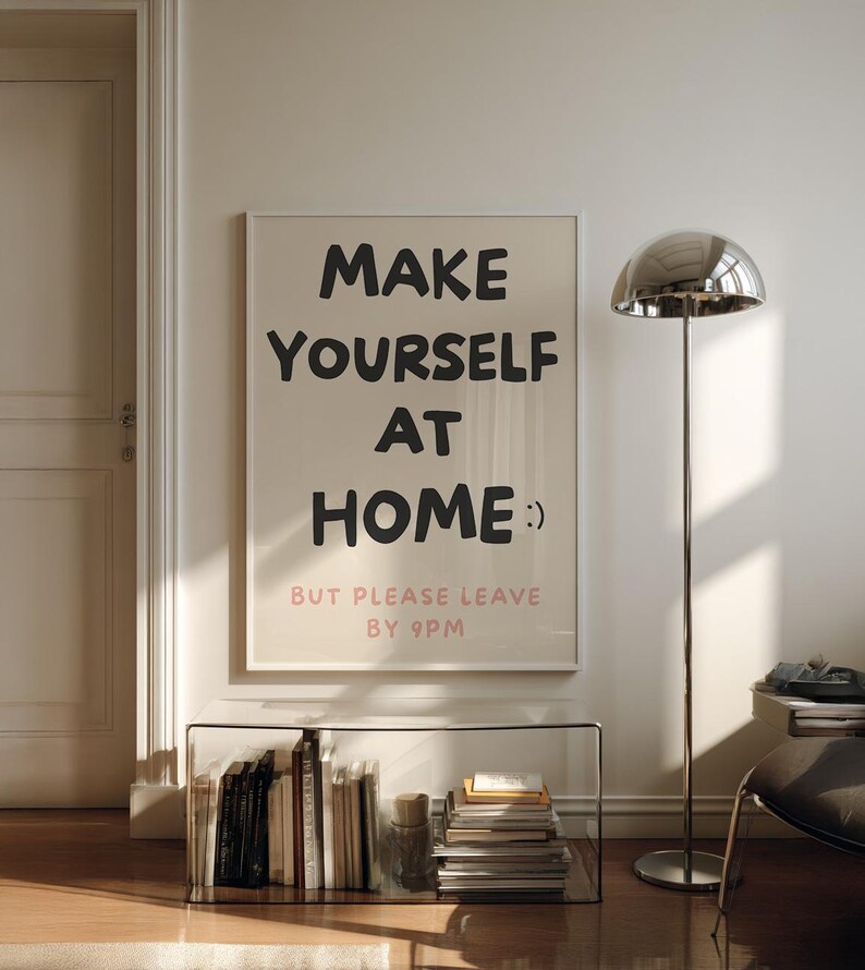 K&ouml;nnte beinhalten: Gerahmtes Poster mit dem Text "MAKE YOURSELF AT HOME :) BUT PLEASE LEAVE BY 9PM" in Schwarz und Rosa. Das Poster h&auml;ngt an einer wei&szlig;en Wand &uuml;ber einem Glasregal mit B&uuml;chern. Rechts steht eine silberne Stehlampe.
