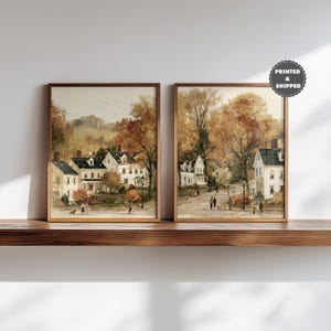 May include: Two framed wall art prints featuring watercolor-style autumn landscapes with white houses, tall trees with orange and brown foliage, and small figures walking along paths. A circular badge in the top right corner reads PRINTED & SHIPPED.