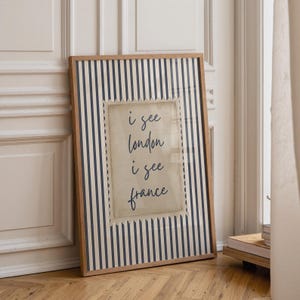 May include: Framed wall art with a wooden frame. The artwork features a striped background in navy blue and white. A central, postage stamp-like design displays the text "i see london i see france" in black script.