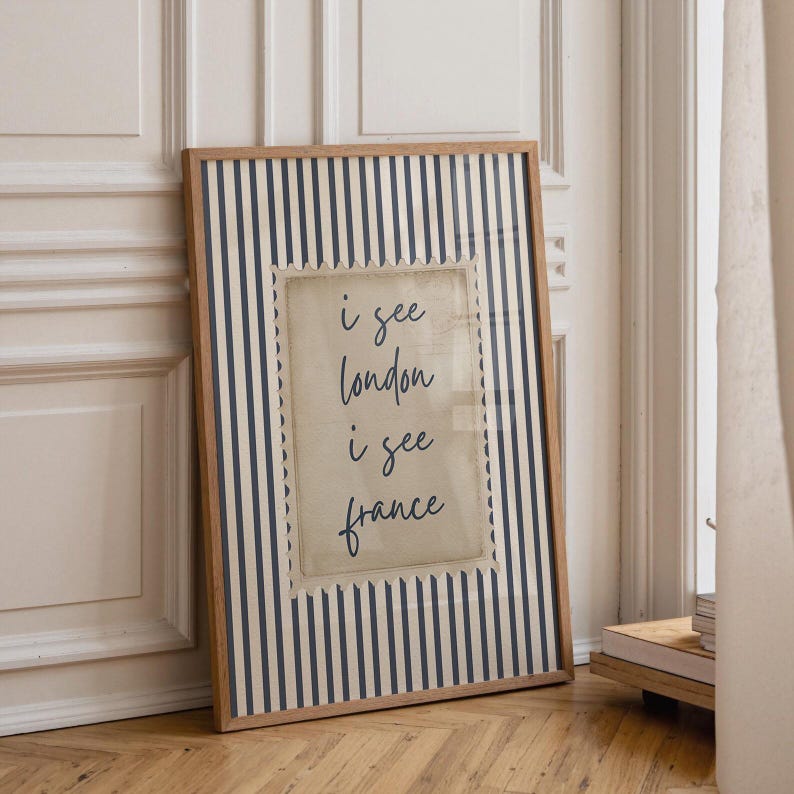May include: Framed wall art with a wooden frame. The artwork features a striped background in navy blue and white. A central, postage stamp-like design displays the text "i see london i see france" in black script.