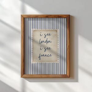 May include: A framed print with a wooden frame. The print features a navy and white striped background with a postage stamp-style image in the center. The text on the stamp reads "i see london i see france".