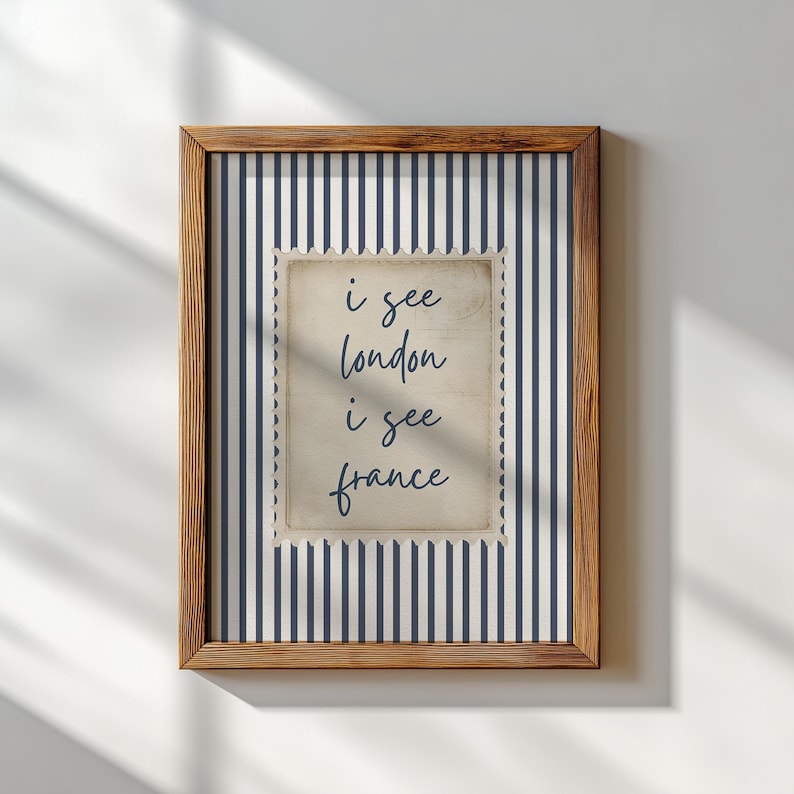 May include: A framed print with a wooden frame. The print features a navy and white striped background with a postage stamp-style image in the center. The text on the stamp reads "i see london i see france".