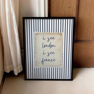 May include: Framed art print with a black border. The background features navy blue and white vertical stripes. A central, postage stamp-shaped design displays the text "i see london i see france" in a handwritten style.