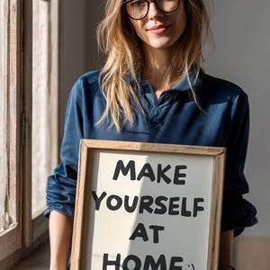 K&ouml;nnte beinhalten: Ein gerahmtes Schild mit der Aufschrift "MAKE YOURSELF AT HOME :) BUT PLEASE LEAVE BY 9PM" in schwarzer und rosa Schrift. Das Schild wird von einer Person gehalten, die ein blaues Hemd und eine Brille tr&auml;gt. Der Hintergrund umfasst ein Fenster und nat&uuml;rliches Licht.