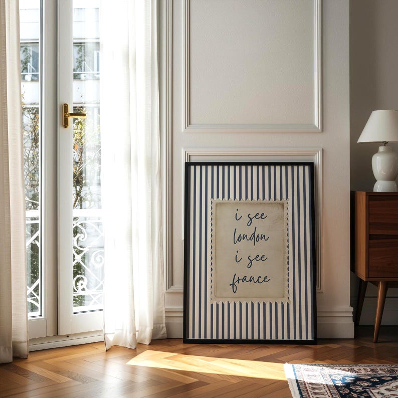May include: A framed print with a black and white striped border. The print features the text "i see london i see france" in a handwritten style. The print is leaning against a white wall next to a window and a wooden cabinet.