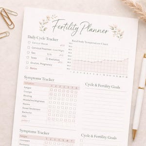 May include: A fertility planner with a floral design, a white pen with gold accents, and gold paper clips. The planner includes sections for tracking daily cycles, symptoms, and fertility goals. The planner is on a light-colored surface.