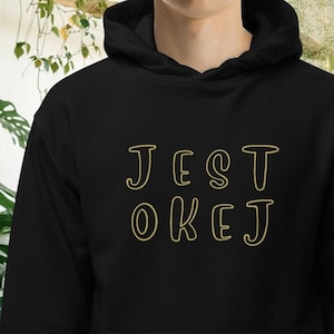 Könnte beinhalten: Schwarzer Kapuzenpullover mit dem Text "JEST OKEJ" in goldfarbenen Buchstaben. Der Pullover hat eine Vordertasche und eine Kapuze mit Kordelzug. Der Hintergrund zeigt grüne Pflanzen.