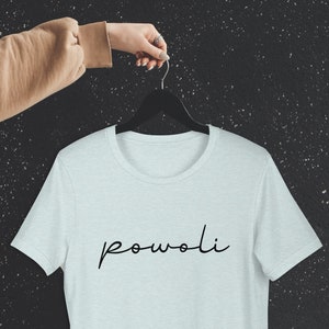 Op de afbeelding: Een lichtblauw t-shirt met het woord "powoli" in zwart script. Het shirt hangt aan een zwarte hanger, vastgehouden door een hand. De achtergrond is een donker, gespikkeld oppervlak.
