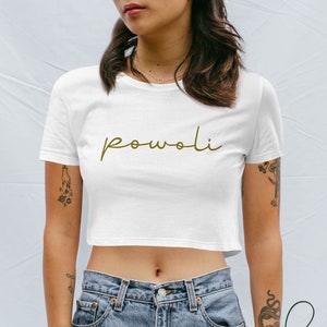 Op de afbeelding: Een witte crop top met het woord "powoli" in gouden script. De top met korte mouwen heeft een ronde hals. Het model draagt een blauwe spijkerbroek.